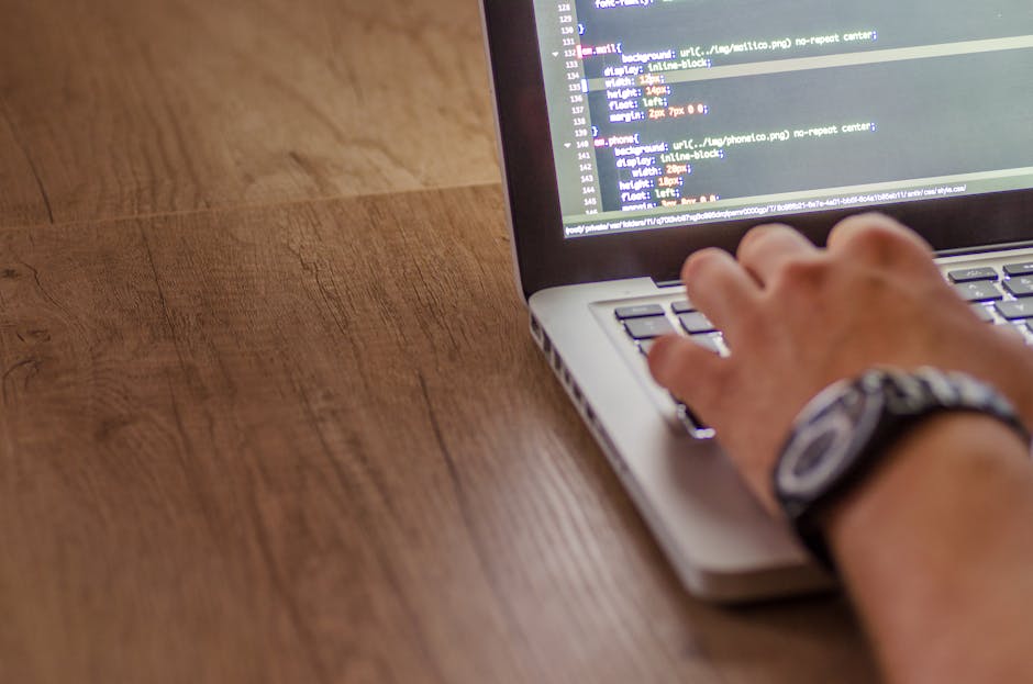 A close-up view of a person's right hand using a laptop placed on a wooden desk, with the screen displaying lines of code in a text editor. The hand is wearing a black and grey patterned wristwatch, a